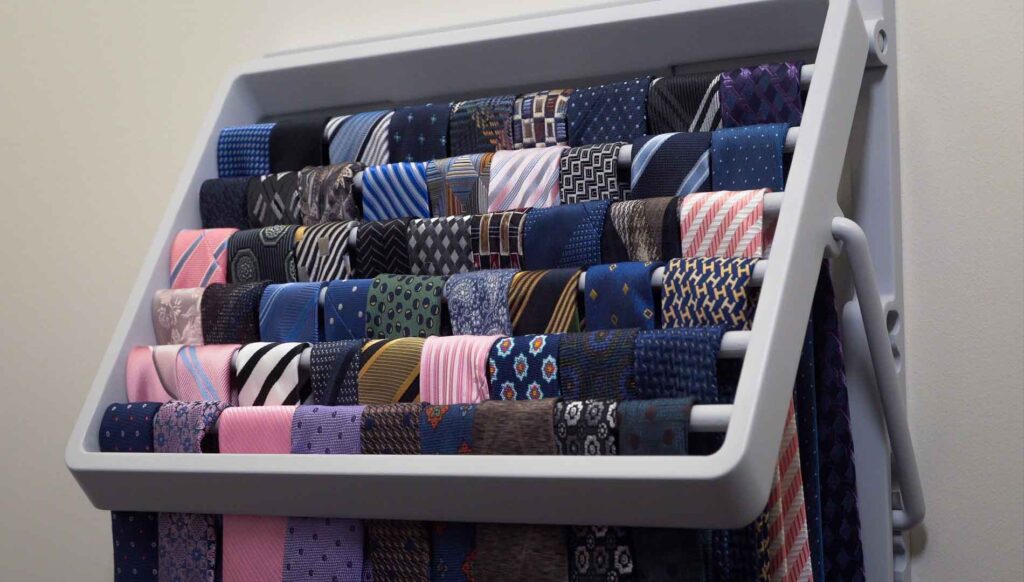 A grey tie rack hangs on a wall. Various colors and patterns of ties hang from its rungs.