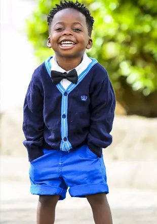 A child wearing a blue cardigan, blue shorts, and a black bowtie smiles broadly at the camera.